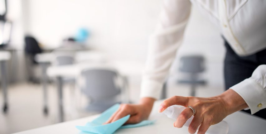 Teacher back at school after covid-19 quarantine and lockdown, disinfecting desks. Unreecognizable teacher back at school after covid-19 quarantine and lockdown, disinfecting desks at break time.